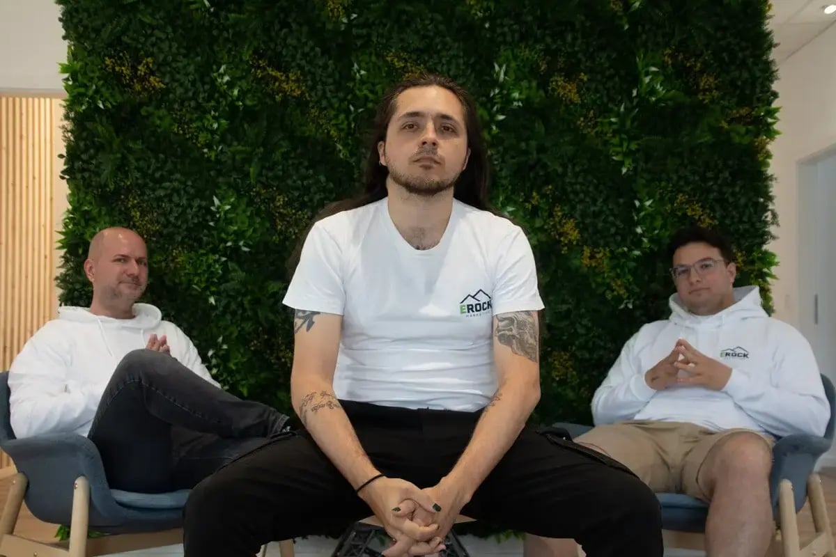 Drei Männer sitzen vor einer grünen Pflanzenwand. Der Mann in der Mitte trägt ein weißes T-Shirt und schaut direkt in die Kamera, während die anderen zwei in weißen Hoodies entspannt im Hintergrund sitzen. Symbolisiert Teamgeist oder ein modernes Arbeitsumfeld.