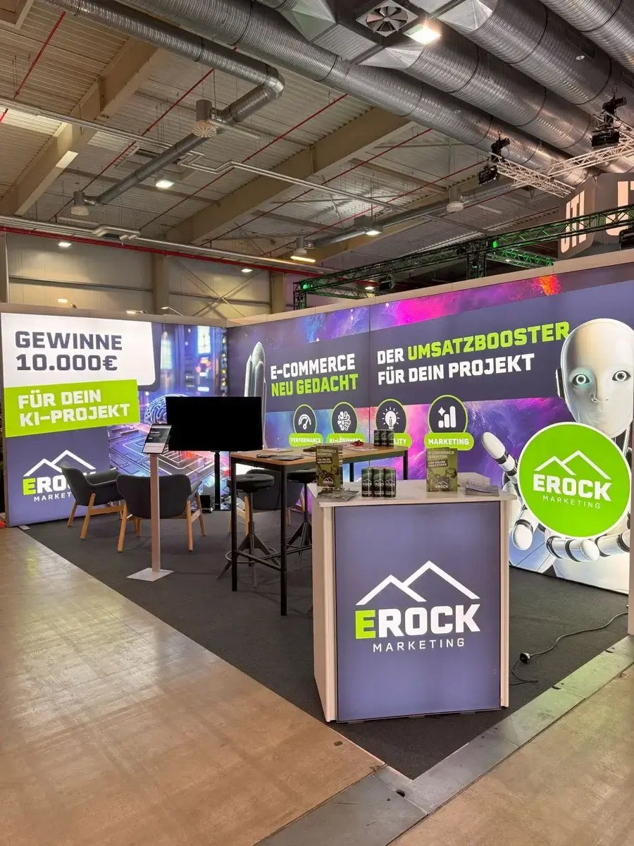 Messestand mit futuristischem Design, großem Bildschirm und Werbebannern. Slogans wie 'E-Commerce neu gedacht' und 'Der Umsatzbooster für dein Projekt'. KI-Roboter-Grafik und 10.000€-Gewinnspiel. Symbolisiert Innovation und Digitalisierung im E-Commerce.