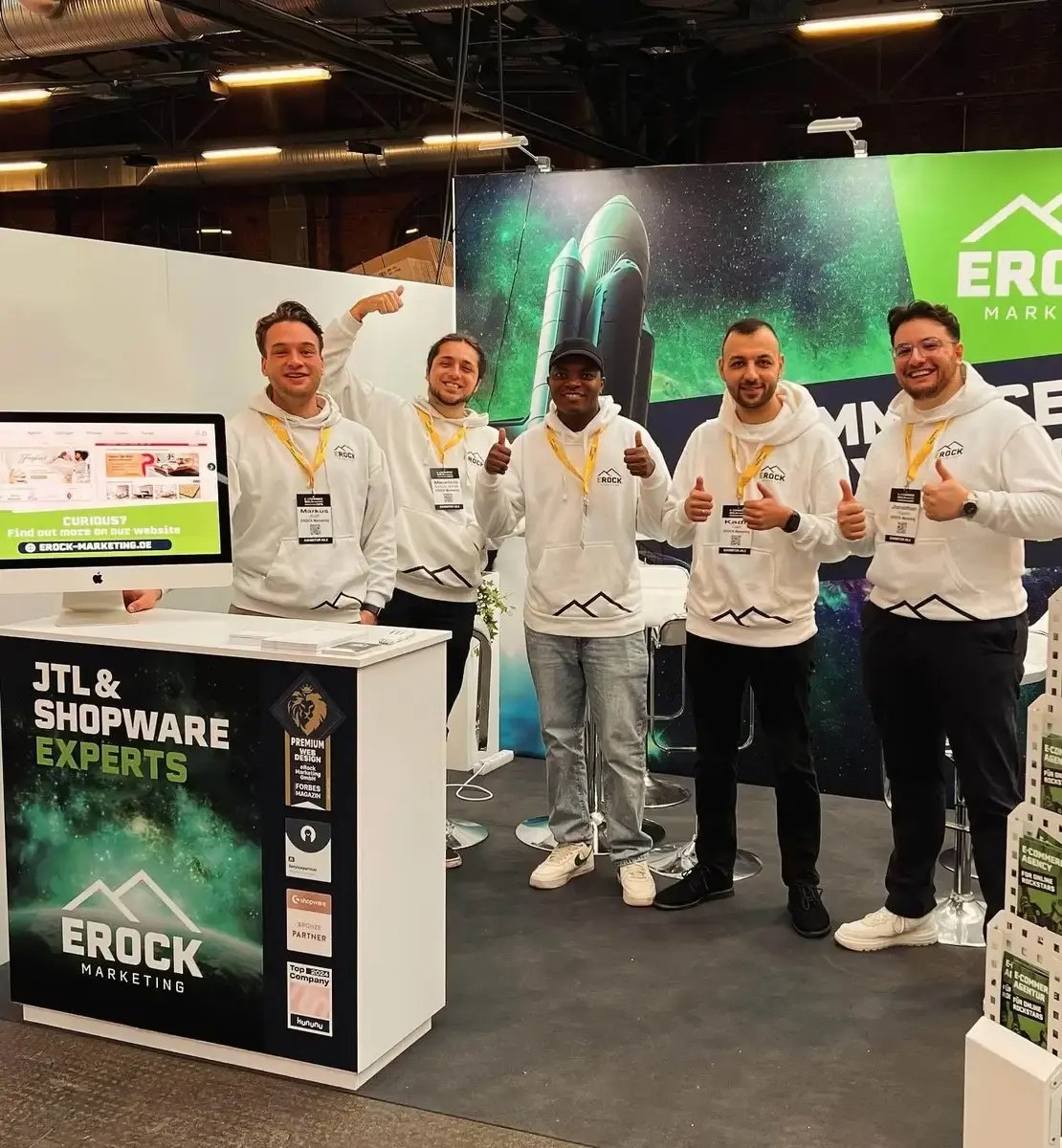Fünf Männer in weißen Hoodies mit Logo stehen an einem Messestand, lächeln und zeigen Daumen hoch. Der Stand wirbt für 'JTL &amp; Shopware Experts' mit einem Monitor und Bannern im Hintergrund. Symbolisiert Teamwork, E-Commerce-Expertise und digitales Business.