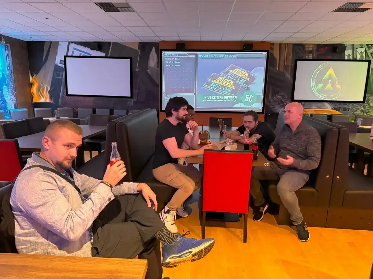 Gruppe von Männern sitzt entspannt in einer Lounge mit Getränken. Im Hintergrund mehrere Bildschirme mit Präsentationen oder Spielen. Symbolisiert Networking, Team-Event oder entspanntes Beisammensein nach einer Messe oder Konferenz.