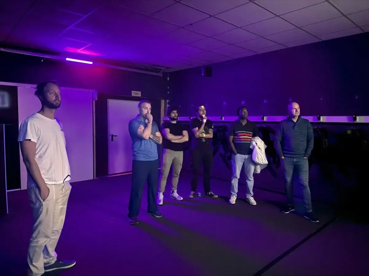 Sechs Männer stehen in einem dunklen Raum mit violetter Beleuchtung. Einige haben die Arme verschränkt, andere halten Jacken oder beobachten etwas. Symbolisiert Team-Event, Gaming-Session oder eine Präsentation in entspannter Atmosphäre.