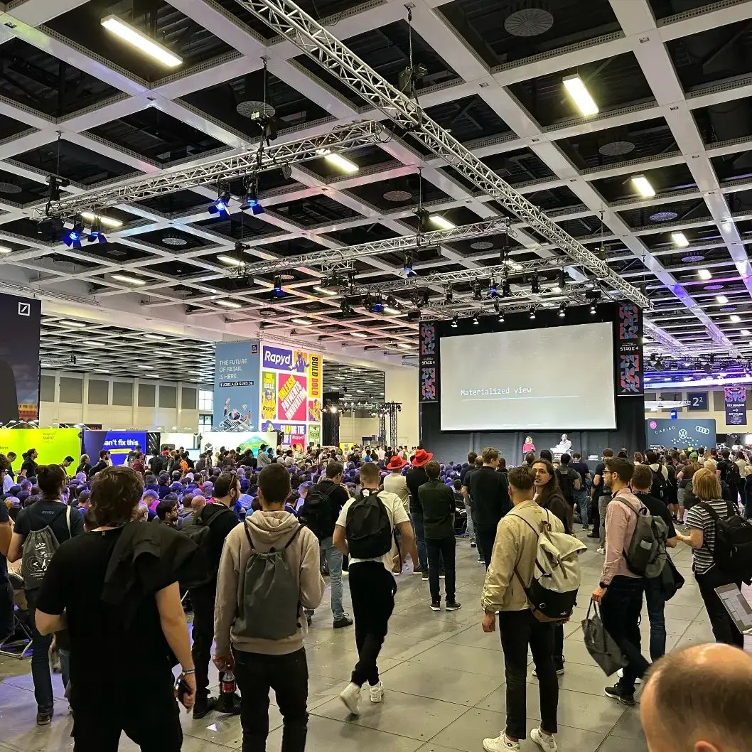 Große Messehalle mit vielen Besuchern, die sich um eine Bühne mit großer Leinwand versammeln. Zahlreiche Stände und Banner im Hintergrund. Symbolisiert eine Fachmesse, Konferenz oder Networking-Event im digitalen oder technologischen Bereich.