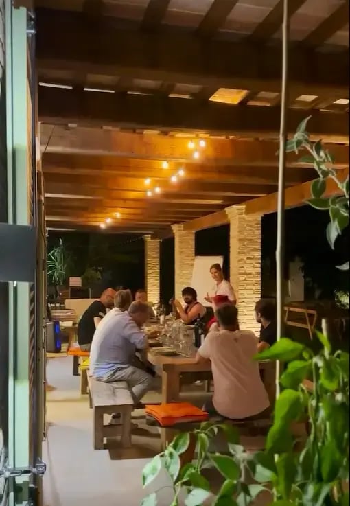 Gruppe von Menschen sitzt an einem langen Holztisch unter einer überdachten Terrasse mit warmen Lichterketten. Eine Person steht und spricht, während die anderen zuhören. Symbolisiert Team-Event, Networking oder gemütliches Beisammensein.