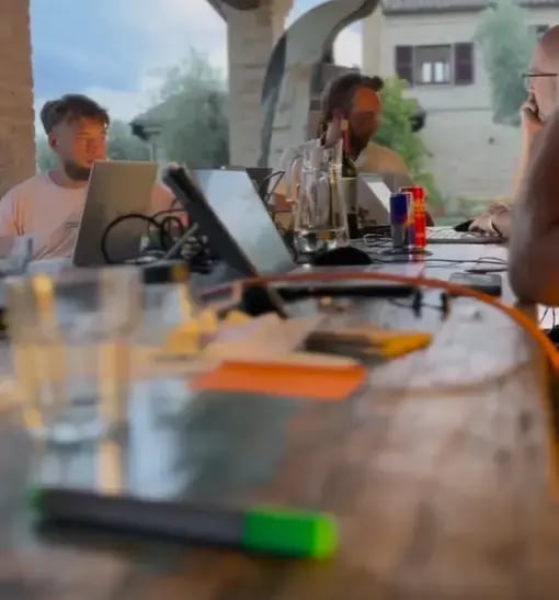 Mehrere Personen sitzen mit Laptops an einem Tisch in einer offenen, mediterranen Umgebung. Getränke und Arbeitsmaterialien liegen auf dem Tisch. Symbolisiert Remote-Work, Team-Meeting oder kreatives Arbeiten im Freien.